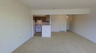 This single-floor space features an open-concept design that connects the living area to the kitchen, creating a functional and cohesive layout. The living area is finished with wall-to-wall neutral-toned carpet, providing a comfortable and unified feel throughout the space. Walls are painted in a clean, light neutral tone, contributing to a bright and airy ambiance.

The kitchen is positioned adjacent to the living area and is defined by dark wood cabinetry that offers ample storage. A white range with overhead ventilation serves as the primary cooking appliance. A white peninsula with a breakfast bar overhang extends into the open living space, providing additional counter space and a casual seating or dining option. The kitchen floor transitions to a hard surface, differentiating it from the carpeted living zone.

A doorway visible toward the rear of the space leads to additional rooms, suggesting a practical and straightforward floor plan. Ventilation registers are visible at the base of the walls, indicating a central heating and cooling system. Overall, the space presents a clean, modest, and functional environment suited for everyday living.