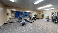 This fitness center offers a comprehensive workout environment designed to accommodate a variety of exercise routines. The space is laid out across a single floor and is anchored by a selection of cardio equipment, including treadmills and elliptical machines, each equipped with individual display consoles for tracking workouts. A multi-station strength training machine and a cable pulley system provide ample options for resistance and weight training, making the facility well-suited for full-body exercise.

The interior is defined by a bold color palette — warm beige walls contrast sharply with a vivid blue accent wall that serves as a striking visual focal point. Full-length mirrors line one wall, enhancing the sense of space and allowing users to monitor their form during exercise. Overhead fluorescent lighting ensures the room is consistently and evenly illuminated throughout.

Durable, patterned carpet tiles cover the floor, offering both comfort underfoot and a polished, functional aesthetic. Potted greenery adds a touch of life to the room, while framed artwork decorates one wall, contributing to a welcoming atmosphere. A small storage shelf provides space for accessories, and a dedicated exit door ensures convenient access. Overall, the fitness center is practical, organized, and thoughtfully appointed.