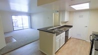 This single-story space showcases an open-concept layout that seamlessly connects the kitchen and living area. The kitchen is anchored by a peninsula-style counter with dark countertops that contrast with crisp white cabinetry, providing both prep space and a natural division between the two areas. The peninsula features a double-basin sink and is complemented by a stainless steel dishwasher and additional upper and lower white cabinets along the perimeter wall, offering generous storage. A skylight above the kitchen introduces natural light, brightening the workspace. The flooring transitions from light hardwood-style material in the kitchen to plush carpeting in the adjoining living area, clearly delineating the two functional zones. The living area benefits from abundant natural light streaming through a large sliding glass door fitted with vertical blinds, which also provides access to an outdoor area. Neutral wall tones throughout the space contribute to a clean, versatile aesthetic. A mirrored accent near the kitchen hallway adds depth to the interior, while a utility or storage closet is accessible nearby, adding to the practical functionality of the layout.