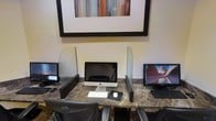 This business center is a single-floor space designed to provide guests or users with a functional and efficient workspace. The room features a continuous marble-patterned desk surface that runs along the wall, accommodating multiple computer workstations side by side. Each station is equipped with a desktop computer — including an Apple iMac and a Samsung monitor — along with keyboards, mice, and mouse pads for convenient use.

Clear acrylic privacy dividers separate each workstation, offering a degree of personal space while maintaining an open feel throughout the room. The ergonomic mesh office chairs at each station enhance comfort during extended use. The warm, neutral-toned walls contribute to a calm and focused atmosphere, while a large framed piece of abstract wall art with warm red, orange, and gray tones adds a decorative touch above the desk. The overall design balances practicality with a polished aesthetic, making it a well-appointed space suited for productivity, whether for checking emails, printing documents, or conducting research.