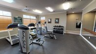 This fitness center is a comprehensive, single-floor facility designed to support a wide range of exercise activities. The space is divided into distinct functional zones, each thoughtfully outfitted to meet various fitness needs.

The main workout area features durable rubber flooring and is equipped with a selection of cardio machines, including treadmills, elliptical trainers, and a stationary bike. A dedicated strength training section houses a multi-function cable machine and a weight bench, while a three-tier dumbbell rack holds a full range of free weights. Framed artwork and a posted fitness center rules sign add character and structure to the space.

Natural light filters in through multiple windows fitted with wood-toned blinds, complementing the overhead fluorescent lighting and ceiling fan fixtures. Crown molding runs along the upper walls, adding a refined architectural touch to the otherwise utilitarian space. The textured ceiling further contributes to the room's clean, well-maintained aesthetic.

A separate mirrored studio area is a standout feature, with floor-to-ceiling mirrors spanning an entire wall and a wooden ballet barre mounted at mid-height. This space also includes exercise mats and step platforms, making it well-suited for stretching, dance, or group fitness activities. Together, the two areas form a versatile and fully functional fitness facility.