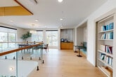 This thoughtfully designed amenity space in Salem, MA combines a café, fitness center, and speakeasy into a single, expansive floor defined by light wide-plank hardwood flooring, crisp white walls, and warm wood accents. The café area features a communal dining setup beneath an elegant chandelier, with large windows offering abundant natural light and views of the surrounding neighborhood. A built-in beverage station with cabinetry adds a functional, polished touch. Adjacent to the café, a cozy nook with tufted banquette seating and tulip side tables invites informal gathering. The lounge area serves as the heart of the space, anchored by a dramatic suspended central fireplace encircled by a curved sectional sofa. Sheer floor-to-ceiling curtains frame panoramic windows, enhancing the bright and airy atmosphere. A dedicated game room features a wooden billiards table beneath a layered brass pendant light. A private media room with a deep-channel sectional sofa and wall-mounted screen provides a more intimate retreat. The fitness center is well-equipped with a range of cardio and strength training machines. A stunning open mezzanine with glass-panel railings, warm wood trim, and sculptural brass pendant lighting completes this sophisticated and versatile amenity floor.