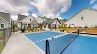 Located at 5150 Adairview Cir in Alpharetta, Georgia, this two-story community amenity space showcases a modern farmhouse aesthetic defined by white board-and-batten siding, a striking metal roof, and barn-style accents. The centerpiece of the outdoor area is a full pickleball court with a vibrant blue surface, enclosed by chain-link fencing and complemented by seating areas for spectators. Adjacent to the court, a meticulously maintained putting green with multiple holes offers residents a dedicated space for golf practice, set against a backdrop of lush, manicured lawn and string lights that create an inviting ambiance for evening gatherings. A covered porch extends from the main structure, furnished with comfortable seating and a mounted screen, making it ideal for outdoor entertaining. The grounds also include raised cedar garden beds set along a gravel pathway, bordered by a classic black horizontal-rail fence and young landscaping that adds greenery and texture to the environment. The surrounding neighborhood of craftsman and cottage-style homes frames the amenity area, contributing to a cohesive, community-focused atmosphere. Together, these features create a well-rounded outdoor destination designed to promote recreation, leisure, and neighborly connection in the heart of Alpharetta.