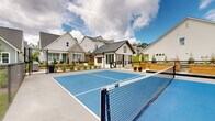 Located at 5150 Adairview Cir in Alpharetta, Georgia, this two-story community amenity space showcases a modern farmhouse aesthetic defined by white board-and-batten siding, a striking metal roof, and barn-style accents. The centerpiece of the outdoor area is a full pickleball court with a vibrant blue surface, enclosed by chain-link fencing and complemented by seating areas for spectators. Adjacent to the court, a meticulously maintained putting green with multiple holes offers residents a dedicated space for golf practice, set against a backdrop of lush, manicured lawn and string lights that create an inviting ambiance for evening gatherings. A covered porch extends from the main structure, furnished with comfortable seating and a mounted screen, making it ideal for outdoor entertaining. The grounds also include raised cedar garden beds set along a gravel pathway, bordered by a classic black horizontal-rail fence and young landscaping that adds greenery and texture to the environment. The surrounding neighborhood of craftsman and cottage-style homes frames the amenity area, contributing to a cohesive, community-focused atmosphere. Together, these features create a well-rounded outdoor destination designed to promote recreation, leisure, and neighborly connection in the heart of Alpharetta.