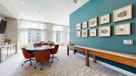 This single-floor amenity space is thoughtfully designed to offer a diverse range of social and recreational functions, all tied together by a cohesive contemporary aesthetic. A consistent patterned carpet in neutral tones, crisp white walls, and bold teal accent walls create visual continuity throughout. Warm cognac leather seating appears across multiple rooms, reinforcing a unified design language.

The game room is a standout area, featuring a full-size pool table bathed in natural light from large windows, a bar-height counter, and teal accent walls adorned with framed artwork. An adjacent conference-style room offers a round table with rolling leather chairs, a shuffleboard table, and a curated gallery wall. The screening room provides rows of cushioned recliner chairs facing a large wall-mounted television, with a popcorn machine adding a cinematic touch.

A bright, open lounge area features multiple seating arrangements and floor-to-ceiling windows with French doors opening to outdoor views. An activity nook houses a puzzle and game table alongside a well-stocked bookshelf. The floor is connected by a wide carpeted corridor with dark wood railings, and a stairwell landing showcases sculptural woven pendant lights as a striking focal point.