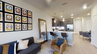 This single-floor residence offers a thoughtfully designed open-concept layout that seamlessly connects the living area and kitchen, creating a bright and functional living environment. The living space features light wood-style flooring throughout, contributing to a warm and modern aesthetic. A charcoal sofa and a tan accent chair are arranged around a small circular side table, complemented by a large floor-length mirror and a bold gallery wall of framed numbered artwork that adds a playful, urban character to the space. The kitchen is a standout feature, showcasing dark wood cabinetry that contrasts elegantly with a crisp white quartz countertop and a breakfast bar with seating for multiple guests. Pendant lighting hangs above the island, providing both task illumination and a refined design touch. Stainless steel appliances, including a refrigerator and microwave, round out the contemporary kitchen. Recessed lighting throughout the space ensures consistent, clean illumination. The overall ambiance is modern and inviting, with a careful balance of dark and light tones, curated furnishings, and clean architectural lines that make this residence both stylish and comfortable for everyday living.