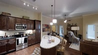 This single-floor residence showcases a thoughtfully designed open-concept layout that seamlessly connects the kitchen, dining, and living areas into one cohesive, flowing space. The kitchen is a standout feature, boasting rich dark-stained cabinetry that extends to the ceiling, stainless steel appliances including a range and built-in microwave, and a striking oval-shaped island topped with light granite countertops. The island doubles as a casual dining spot with seating for multiple guests, making it ideal for both meal prep and entertaining. Elegant pendant lighting hangs above the island, complementing the track lighting overhead and adding warm, layered illumination throughout the space. Crown molding along the ceiling lends a refined architectural touch, while dark hardwood-style flooring grounds the interior with a sense of warmth and continuity. The open living area beyond features a ceiling fan and easy access to adjacent rooms, maintaining an airy, connected feel throughout. Natural light filters in from a nearby doorway, brightening the space and enhancing its welcoming ambiance. Overall, this residence offers a well-balanced blend of functional design and stylish finishes.