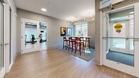 This single-floor space is designed as a communal amenity area, combining a welcoming entryway, a casual lounge, and a dedicated fitness room within a clean, contemporary interior. The entry features an automatic sliding glass door with a transom window above, allowing natural light to filter in while providing a secure, accessible point of entry. The flooring throughout the main area consists of light hardwood-style planks that lend warmth to the otherwise neutral palette of soft gray and beige walls. Recessed ceiling lights and a modern multi-arm pendant fixture illuminate the lounge area, where a tall communal table surrounded by rich wood bar-height chairs sits atop a textured area rug. Colorful abstract artwork adorns the walls, adding visual interest to the space. Large windows along one wall frame views of the surrounding exterior, enhancing the sense of openness. An adjoining room, visible through glass-paneled doors, serves as a fitness area equipped with exercise machines and additional flooring. A secondary, more minimal room with gray walls and light wood flooring offers a versatile, flexible-use space. Together, these areas create a functional and well-appointed communal environment.