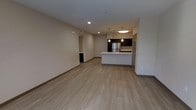 This single-floor unit offers a clean, contemporary design centered around an open-concept living and kitchen area. The kitchen is a standout feature, outfitted with dark espresso cabinetry that contrasts sharply against bright white countertops and a large peninsula island with an integrated sink. Stainless steel appliances, including a full-size refrigerator, add a polished, modern finish. Pendant lighting hangs above the island, complemented by recessed ceiling fixtures and a flush-mount light that ensure the space is well-lit throughout. Light wood-look plank flooring extends seamlessly across the entire main living area, lending warmth and continuity to the open floor plan. The living area is generously proportioned, offering flexible space for a variety of furniture arrangements. Neutral, light-toned walls throughout create a bright and airy ambiance, enhancing the sense of openness. A hallway visible from the main living space provides access to additional rooms. The overall aesthetic is modern and understated, with a cohesive design palette of dark cabinetry, white surfaces, and warm flooring that gives the unit a move-in-ready, well-appointed feel.
