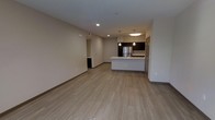 This single-floor unit offers a clean, contemporary design centered around an open-concept living and kitchen area. The kitchen is a standout feature, outfitted with dark espresso cabinetry that contrasts sharply against bright white countertops and a large peninsula island with an integrated sink. Stainless steel appliances, including a full-size refrigerator, add a polished, modern finish. Pendant lighting hangs above the island, complemented by recessed ceiling fixtures and a flush-mount light that ensure the space is well-lit throughout. Light wood-look plank flooring extends seamlessly across the entire main living area, lending warmth and continuity to the open floor plan. The living area is generously proportioned, offering flexible space for a variety of furniture arrangements. Neutral, light-toned walls throughout create a bright and airy ambiance, enhancing the sense of openness. A hallway visible from the main living space provides access to additional rooms. The overall aesthetic is modern and understated, with a cohesive design palette of dark cabinetry, white surfaces, and warm flooring that gives the unit a move-in-ready, well-appointed feel.