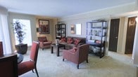 This single-floor space presents a thoughtfully designed living area that combines comfort and style in equal measure. The lounge is anchored by a plush, dark-toned sofa adorned with bold red and white patterned throw pillows, complemented by a set of striped red and cream accent chairs that add a vibrant pop of color. A dark wood coffee table sits at the center, grounding the seating arrangement and providing a functional focal point for the room. Soft, neutral-toned carpeting runs throughout, contributing to a warm and inviting ambiance. The walls are finished in a muted taupe, offering a calm backdrop that allows the furnishings and decor to stand out. A framed piece of wall art in rich reds and blacks adds a decorative focal point, while open black shelving units on either side of the room provide display space for decorative vases, books, and curated accessories. Natural light filters into the space through a window, brightening the room and enhancing its airy feel. Crown molding along the ceiling adds a refined architectural detail, elevating the overall aesthetic. The layout is open and well-organized, making efficient use of the available space.