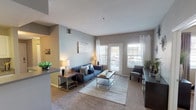 This single-story residence offers a well-organized layout across a comfortable footprint. The living room serves as the central gathering space, finished with plush carpet, neutral gray walls, and large windows alongside a glass door that invite natural light and provide outdoor views. Sheer white curtains complement the bright, open feel of the room.

The galley-style kitchen runs parallel to the living area and features wood-look flooring, white cabinetry, a dual-basin stainless steel sink, and stainless steel appliances including an electric range and refrigerator. A raised countertop peninsula separates the kitchen from the adjacent living space, offering a casual bar-style seating option.

The primary bedroom is carpeted and well-proportioned, with a large window dressed in layered curtains providing ample natural light. The second bedroom, also carpeted, functions well as a guest room or flexible space, featuring its own window with views to the exterior.

Both bathrooms maintain a consistent design with gray walls, wood-look flooring, white vanity cabinetry, and tub-shower combinations with tiled surrounds. The overall aesthetic throughout the unit is cohesive, favoring a modern neutral palette with gray and white tones.