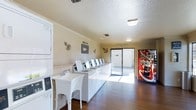 This single-floor communal laundry facility is designed for practicality and ease of use. A row of top-loading washing machines lines the central counter, accompanied by stacked dryer units along the wall, providing ample capacity for multiple users. A standalone utility sink offers added convenience for hand-washing or pre-treating garments. The room features warm hardwood-style flooring that contrasts with the bright white appliances and cream-toned walls, creating a clean and welcoming environment. A textured ceiling with flush-mounted lighting ensures the space is well-lit throughout. Decorative nautical-themed wall accents add a touch of character to the otherwise functional room. A Coca-Cola vending machine is positioned near the entrance, offering refreshments during laundry sessions. At the far end, a large glass sliding door floods the room with natural light and provides direct outdoor access, enhancing the open feel of the space. Crown molding along the upper walls adds a subtle architectural detail. Overall, this laundry room is thoughtfully arranged to balance functionality with a comfortable, tidy atmosphere.