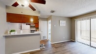 Located at 6701 Everhart Rd in Corpus Christi, Texas, this single-floor, one-bedroom, one-bathroom unit offers a practical and well-organized layout. The living room features wood-look laminate flooring, neutral warm-toned walls, a ceiling fan, and a sliding glass door fitted with vertical blinds that open to a balcony, allowing natural light to fill the space. The open-concept design connects the living area directly to the kitchen, which is separated by a half-wall breakfast bar. The kitchen is equipped with warm wood cabinetry, a coil-burner electric range with overhead ventilation hood, and light-speckled countertops. The bedroom is generously sized relative to the overall footprint, finished with plush carpet flooring, a large window with exterior views of mature trees, and an expansive built-in closet with full-panel sliding doors offering substantial storage. The bathroom includes a full soaking tub and shower combination with clean white surrounds and wood-look flooring. A textured popcorn ceiling is consistent throughout the unit, complementing the understated, neutral color palette. The exterior reflects a traditional multi-unit building with siding construction and mature tree coverage.