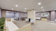 The Resident Lounge at 8040 Rowland Avenue in Philadelphia, PA offers a well-appointed, single-floor community gathering space designed with both comfort and social engagement in mind. The lounge is finished throughout with soft, neutral carpeting and warm mauve-toned walls, creating a cohesive and inviting ambiance accented by recessed lighting overhead.

The main lounge area is anchored by a central fireplace framed in light wood, surrounded by modern white armchairs that invite relaxed conversation. Adjacent to this focal point, a contemporary sofa arrangement is complemented by a natural wood planter box filled with lush greenery, adding a refreshing organic touch. A curated gallery wall and a large statement clock further enhance the room's character.

A dedicated recreation zone features a shuffleboard table alongside additional casual seating, perfect for social gatherings and recreation. A built-in coffee bar with open shelving provides a convenient amenity for residents. 

Transparency and openness are emphasized through floor-to-ceiling glass partitions that visually connect distinct areas of the lounge, including a separate multipurpose room with hardwood flooring, wall murals, and a wall-mounted split system for climate control. A long built-in bench with integrated planters lines one wall near the elevator, offering a welcoming transition space. The lounge is thoughtfully designed to serve as a true community hub for residents.