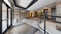 Located at 3777 Rue St-Urbain in the heart of Montréal, this building lobby serves as the central ground-floor entrance and circulation hub for the property. The space is defined by its distinctive granite-clad walls accented with warm terracotta-toned horizontal banding, lending a classic and polished character throughout.

Upon entry, floor-to-ceiling glass doors and sidelights flood the vestibule with natural light, offering clear sightlines to the exterior. The main lobby area features recessed ceiling lighting set within a coffered ceiling detail with dark trim, adding architectural depth overhead. Smooth tile flooring transitions seamlessly across the space, complemented by low-pile entry mats at thresholds.

A curved staircase with sleek black metal railings leads to the raised main hallway level, which also offers an accessible ramp route. Two elevators are prominently positioned along the granite feature wall, labeled for ground-floor access. Adjacent to the elevator bank, a built-in mailbox unit serves residents efficiently.

The hallway extends from the lobby, lined with granite walls and recessed lighting, creating a clean and functional corridor. Potted bamboo and leafy green plants are strategically placed throughout, softening the hardscape materials and adding a touch of life and warmth. Overall, the lobby presents a well-maintained, functional, and professionally finished communal space.