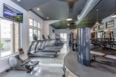 This single-level fitness center is thoughtfully designed to accommodate a wide range of workout needs within a spacious, open layout. The entry area features polished concrete floors, a storage credenza with cubbies, and a wall-mounted AED unit, setting a clean and safety-conscious tone. A boldly striped green and white accent wall adds a pop of color near the entrance.

The main workout floor is divided into distinct zones. The cardio and strength training area boasts soaring ceilings with exposed dark ductwork, pendant lighting, and large windows that invite natural light and outdoor views. The space is well-appointed with treadmills, cable machines, rowing equipment, adjustable benches, and a full dumbbell rack.

A dedicated functional training zone features polished concrete flooring, a battle rope suspended from the ceiling, a speed bag, a clothing rack, and a striking large-scale mural of a runner rendered in a particle-dispersion style—lending an energetic, motivational atmosphere. Rubber sport flooring with yellow border accents defines the training platform area. Throughout, the industrial-modern aesthetic blends dark ceiling tones with light walls and reflective flooring for a dynamic, high-energy environment.