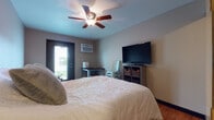 Located at 1905 Miller St in La Crosse, Wisconsin, this single-floor studio offers a well-organized and efficiently designed living space. The main area serves a dual purpose, combining a sleeping zone and a relaxing sitting nook. The sleeping area features hardwood flooring, a ceiling fan with integrated light fixture, a wall-mounted air conditioning unit, and a private exterior door that allows natural light to filter in. A wall-mounted flat-screen TV and compact storage shelving complete the space. Adjacent to the sleeping area, a cozy sitting nook is furnished with a sectional sofa, a plush area rug, and its own exterior door, creating a bright and comfortable retreat. A sliding barn-style door provides a stylish transition between the living areas. The kitchenette is equipped with a rustic wood-top counter, a toaster oven, and a gas cooktop, offering essential cooking convenience. The bathroom includes hardwood flooring, warm wood cabinetry for overhead storage, and a shower area. Throughout the studio, warm-toned walls, consistent hardwood flooring, and practical design choices create a cohesive and inviting atmosphere suited for comfortable everyday living.