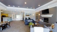 This single-story community space offers a thoughtfully designed interior that balances comfort and functionality. The main lounge area is anchored by a classic white-mantled fireplace set against a warm accent wall, flanked by built-in shelving displaying potted greenery. A wall-mounted television above the fireplace adds a modern touch. Plush navy blue sofas and accent chairs are arranged around a wood-and-metal coffee table, all resting on patterned carpeting that adds texture to the space.

A tray ceiling with recessed lighting and crown molding lends architectural distinction to the room, while wall sconces provide soft, ambient illumination throughout. A dedicated workstation nook, tucked into a recessed alcove and lit by pendant lights, provides a quiet area equipped with desktop computers — ideal for residents or guests needing a productive workspace.

Large French doors along the far wall flood the interior with natural light and provide a welcoming connection to the outdoors. The overall color palette of grays, warm taupes, and navy blues creates a cohesive, inviting ambiance suited for both socializing and focused work. The space is clean, modern, and well-maintained, reflecting a high standard of community amenity design.
