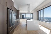 This contemporary high-rise residence offers a refined living experience defined by clean lines, premium finishes, and sweeping waterfront views visible from multiple rooms. Light hardwood flooring flows throughout the open layout, complementing the neutral palette of soft whites and warm grays that lend the space a calm, sophisticated ambiance.

The chef's kitchen is a standout feature, showcasing floor-to-ceiling cabinetry in a rich wood-grain finish, a generous quartz waterfall island with an integrated gas cooktop, a stainless steel range hood, double wall ovens, a panel-ready refrigerator, and a built-in wine cooler — all designed for both function and style.

The living areas benefit from expansive floor-to-ceiling windows that frame breathtaking panoramic views of the bay and surrounding cityscape, flooding the rooms with abundant natural light. A wide, well-lit corridor connects the private quarters, which include spacious bedrooms with large windows capturing the same striking views. The walk-in closet is outfitted with custom dark wood shelving and hanging systems, providing generous and well-organized storage.

The bathrooms are finished with large-format tile flooring, floating wood vanities, illuminated mirrors, and glass-enclosed walk-in showers, maintaining the home's cohesive modern aesthetic throughout.