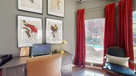 This thoughtfully designed single-floor amenity space combines functionality and refined aesthetics in a welcoming environment. The interior is anchored by a well-equipped business center, featuring a large gray wood-tone desk with built-in storage drawers, a leather executive chair, and a brass accent lamp — ideal for residents needing a productive workspace. Large grid-pane windows dressed with bold crimson drapes frame views of a brick-clad courtyard and sparkling pool, flooding the space with generous natural light throughout the day.

The walls are finished in a muted sage-gray tone that complements the warm hardwood-style flooring, creating a cohesive and polished atmosphere. A curated gallery wall of framed botanical bird illustrations adds an artistic, sophisticated touch to the workspace area.

A comfortable lounge nook anchors another corner of the space, furnished with a pair of ornate French-style chairs upholstered in dark leather, positioned beside a rustic wood side table — perfect for informal conversation or relaxed reading.

Elegant white French doors with glass pane inserts connect the main area to additional amenities, including a fitness center visible through a glass partition. The overall ambiance is warm, curated, and inviting, blending classic design elements with practical community amenities.