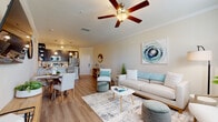 This single-floor residence offers a thoughtfully designed open-concept layout that seamlessly connects the kitchen, dining, and living areas. The living room is bright and welcoming, featuring crown molding, a ceiling fan, light wood-style flooring, and multiple windows dressed with teal curtains that bring in generous natural light. The space is well-proportioned, providing ample room for comfortable seating and entertaining.

The kitchen is outfitted with dark espresso cabinetry, stainless steel Samsung appliances — including a refrigerator, range, and microwave — and a light quartz countertop with a peninsula that doubles as a breakfast bar with pendant lighting above. Crown molding continues throughout, reinforcing the polished aesthetic.

The dining area sits adjacent to the kitchen and living room, highlighted by an arched architectural detail that adds a classic touch. The bedrooms feature plush carpeting and generous proportions, with one bedroom offering a walk-in closet. The bathroom includes a full soaking tub and shower combination with tiled surround, dark vanity cabinetry, and wood-style flooring.

A dedicated laundry room houses a full-size Samsung washer and dryer with overhead shelving for added storage. Throughout the residence, the neutral warm palette and coastal-inspired décor create a calm, cohesive ambiance.
