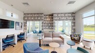Located at 1708 Starship Dr in Cocoa, Florida, this single-floor community space offers a bright, contemporary design across a well-utilized footprint. The interior is finished with light hardwood flooring throughout and painted in soft gray tones, creating a clean and welcoming atmosphere.

The two living areas serve distinct purposes. The main lounge is generously furnished with a plush gray sofa and modern accent chairs in blue and teal, centered around a compact side table. Large floor-to-ceiling windows and glass doors flood the space with natural light while offering direct views of the surrounding green landscape and outdoor amenity areas. A secondary lounge zone features a built-in workstation with two chairs, a wall-mounted television, pendant lighting, and a glass partition wall that subtly divides the space.

The dining area is enclosed with glass panel walls and showcases a round wood-top table surrounded by vibrant blue chairs. A wall-mounted flat-screen television and a tall window framing pond and nature views complete this intimate setting.

Recessed lighting throughout ensures consistent illumination, while multiple entry points with glass doors connect the interior to the outdoors. The overall ambiance is modern, airy, and functional — ideal as a shared gathering and working space.