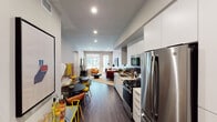 This contemporary single-floor residence offers a thoughtfully designed open-concept layout that seamlessly connects the kitchen, dining, and living areas. The galley-style kitchen is outfitted with sleek white cabinetry extending to the ceiling, a dark tile backsplash, light quartz countertops, and a full suite of stainless steel appliances including a gas range, built-in microwave, dishwasher, and French door refrigerator. Rich dark hardwood-style flooring runs throughout the main living areas, grounding the clean, modern aesthetic. The dining nook flows naturally into the living room, which benefits from large windows and a glass door that invite abundant natural light and provide views of the surrounding streetscape. The living room offers a comfortable, versatile layout with wall space for a large-format television. The bedroom features a ceiling fan, a generous window with outdoor views, and the same durable flooring. A full bathroom includes a soaking tub with shower combination, a vanity with ample storage, and built-in shelving. Recessed lighting throughout the space ensures a bright, welcoming ambiance, while the neutral palette and modern finishes create a cohesive and contemporary feel.