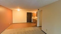 This single-floor residential space offers a straightforward and functional layout designed for comfortable everyday living. The main living area is defined by a combination of smooth hardwood-style flooring and plush carpeting, creating distinct zones for movement and relaxation. Warm, neutral-toned walls contribute to an inviting and cozy ambiance throughout.

The kitchen is open and accessible from the main living space, featuring classic wood-finish upper and lower cabinetry that provides ample storage. A light-toned countertop runs along the workspace, and the layout allows for efficient use of the area. The kitchen and living spaces flow naturally into one another, making the overall footprint feel connected and cohesive.

The main entryway is marked by a sturdy dark-finished door, offering a clear and practical point of entry. Overhead lighting illuminates the space with a warm glow, complementing the earthy tones found throughout the unit. Textured ceilings add a subtle architectural detail to the interior. Ventilation is provided through ceiling-mounted registers, ensuring a consistent and comfortable indoor environment. Overall, the space presents a clean, move-in-ready interior with a warm and practical character suited for daily residential use.