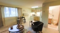 This single-floor unit offers an open-concept design that combines the living and kitchen areas into one cohesive, easy-to-navigate space. The living area is finished with neutral carpet flooring and features soft, light green walls that lend a calm and welcoming ambiance throughout. A window dressed with flowing curtains allows natural light to filter into the room, brightening the space and offering a view to the outside. A wall-mounted HVAC unit provides convenient climate control directly within the living area.

The kitchen is positioned at the rear of the open floor plan and is outfitted with light wood cabinetry, a white refrigerator, a white range with oven, and a sink with overhead task lighting provided by a fluorescent ceiling fixture. The cabinetry offers ample storage, and the layout keeps essentials within easy reach.

The living area comfortably accommodates a seating arrangement, including a cushioned accent chair and a round coffee table, alongside a tall bistro-style table with stools near the window — ideal for casual dining or additional seating. The overall layout is efficient and thoughtfully arranged, making practical use of the available space while maintaining an open, airy feel.