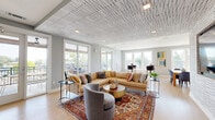 Located at 235 Verbena St in Charlotte, NC, this thoughtfully designed single-floor communal space blends modern aesthetics with functional living areas. The living room is an immediate standout, anchored by a striking geometric patterned ceiling with recessed lighting that adds visual depth and dimension. Light hardwood flooring runs throughout, complementing the soft gray walls and creating a warm, cohesive atmosphere. Large sliding glass doors and multiple windows flood the space with natural light and provide direct access to a covered outdoor terrace, where wood-paneled ceilings, a ceiling fan, and open railings frame sweeping views of the Charlotte skyline and surrounding neighborhood. Back inside, a textured white brick accent wall in the living area houses a wall-mounted flat-screen TV, while curated artwork and stylish furnishings complete the inviting lounge setting. The dining area transitions seamlessly from the living space and features a large communal wood dining table alongside bar-height seating. A well-appointed kitchen with teal cabinetry, open shelving, stainless steel appliances, and white countertops adds both function and a bold pop of color. Multiple windows in the dining area ensure the room remains bright and airy throughout the day.