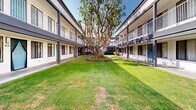 Located in Solana Beach, California, this inviting courtyard serves as the central outdoor hub of a two-story building complex. The space is defined by a generous, well-maintained lawn that stretches along the interior of the complex, anchored by a striking mature tree with a broad, sculptural canopy that provides natural shade and a focal point for the entire area.

Flanking the lawn on both sides are covered walkways lined with unit entrances featuring teal-accented doors, adding a pop of color against the crisp white stucco exterior. Upper-level balconies with metal railings run the full length of both building wings, offering elevated outdoor space for residents and reinforcing the open, airy character of the courtyard.

A dedicated patio area is furnished with wicker lounge chairs and a sofa, all upholstered in bold red cushions, arranged around a central fire pit table. A large red market umbrella provides shade, and a colorful area rug ties the seating arrangement together, creating a warm and social outdoor living environment.

Abundant natural light floods the courtyard throughout the day, reflecting the classic Southern California climate of Solana Beach. Lush planters and shrubs along the building perimeter add greenery and texture, making this courtyard a functional and aesthetically appealing communal outdoor space.