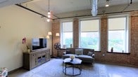 This single-floor Dallas space blends industrial character with comfortable living. The main living area is defined by striking exposed brick walls that span floor to ceiling, lending a warm, textured backdrop throughout. Soaring ceilings reveal architectural elements typical of converted industrial spaces, including visible ductwork and black pipe details, adding an authentic urban aesthetic. Pendant bulb lighting hangs from above, complementing the loft-like atmosphere.

Large, double-hung windows with roller shades line the brick walls, flooding the space with abundant natural light while offering pleasant views of mature trees and the surrounding Dallas cityscape. The generous living area comfortably accommodates a plush sofa, a wood-and-media console, and a layered area rug anchoring the seating arrangement around a modern hexagonal coffee table.

The overall ambiance balances raw industrial charm with welcoming, livable design. The combination of natural light, exposed brick, and carefully chosen furnishings creates a space that feels both spacious and grounded, making it well-suited for both relaxation and everyday living in the heart of Dallas.