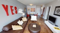 This Dallas apartment showcases a thoughtfully designed open-concept layout, seamlessly blending the living and dining areas. The space is characterized by its modern aesthetic, featuring a polished concrete floor that adds an industrial touch while maintaining a sleek appearance. The living area is anchored by a luxurious brown leather sofa, complemented by a white accent chair and a large circular ottoman, creating a cozy seating arrangement. A striking chrome arc floor lamp serves as both a functional and artistic element.

The dining area, positioned adjacent to the living space, features a dark wood table paired with vibrant red chairs, adding a pop of color to the neutral palette. The walls are painted in a soothing gray tone, providing a perfect backdrop for the eclectic decor, including a large red letter "S" and a round mirror that enhances the sense of space. A contemporary abstract artwork adorns the wall near the entrance, injecting personality into the room.

Storage solutions are seamlessly integrated, with a coat rack visible near the entryway. The apartment's design maximizes natural light, creating an inviting and airy atmosphere. This thoughtfully curated space exemplifies urban living, balancing style and functionality in a compact yet open environment.