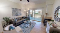This well-appointed single-floor residence offers a thoughtfully designed living space that balances comfort and modern style. The living room serves as the heart of the home, showcasing vaulted ceilings that lend an airy, expansive feel to the interior. Light-toned tile flooring runs throughout, reflecting the abundant natural light that pours in through large sliding glass doors. These doors open directly onto a private patio, creating a seamless indoor-outdoor connection and offering pleasant views of the surrounding exterior. A ceiling fan is centrally positioned to enhance airflow and comfort year-round. The living area is anchored by a tufted charcoal sofa accented with blue and white decorative pillows, complemented by a sculptural glass-top coffee table and a plush area rug in cool blue tones. A statement arc floor lamp and pendant lighting add layered illumination to the space. A bold botanical wall art piece adds a decorative focal point, while the neutral wall tones keep the atmosphere calm and inviting. A fireplace with a light stone surround is visible along one wall, adding a classic architectural element. Overall, the space presents a clean, contemporary aesthetic with warm, welcoming touches throughout.