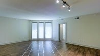 Located at 935 Garden Park Dr. in Allen, Texas, this single-floor space offers a clean, modern aesthetic designed for both comfort and functionality. The main living area is bright and open, characterized by rich hardwood-style flooring that runs throughout and crisp white walls that enhance the sense of openness. Large floor-to-ceiling windows dressed with horizontal blinds allow generous amounts of natural light to pour into the space, creating an airy and welcoming atmosphere. Adjustable track lighting mounted on the ceiling provides versatile, directional illumination to complement the natural light. Crown molding along the ceiling adds a subtle architectural detail that lends the space a polished, refined character. A doorway visible from the main room leads to an additional area, suggesting a practical and well-organized layout. HVAC vents are thoughtfully integrated into the ceiling, ensuring year-round comfort. The overall design reflects a contemporary, minimalist sensibility — clean lines, neutral tones, and quality finishes come together to create a space that is both functional and visually appealing, situated in the thriving community of Allen, TX.
