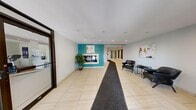 This single-floor lobby space is designed with a clean, contemporary aesthetic that creates a welcoming first impression. The layout consists of a spacious living area connected to an extended hallway, all finished with large-format light-toned ceramic tiles throughout. Recessed lighting illuminates the space evenly, complementing the neutral white and grey walls.

The living area serves as the central gathering point of the lobby, featuring a bold teal accent wall that anchors the room visually. A framed pass-through window is set into this accent wall, adding architectural interest. The seating area includes a pair of sleek black leather chairs arranged around a minimalist side table, accompanied by contemporary abstract artwork on the walls. A potted fiddle-leaf fig plant adds a natural touch near the accent wall.

The hallway extends from the lobby, lined with dark wood-paneled mailboxes and unit doors with grey frames. Security cameras are mounted at the ceiling, and exit signage is clearly visible. At the opposite end, glass entry doors allow natural light to filter into the corridor, offering views to the exterior. The overall ambiance is modern, functional, and well-maintained, with a professional character suitable for a multi-unit residential building.
