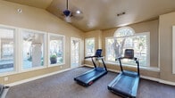 The fitness center at 6111 N Beach St in Fort Worth, Texas, offers a well-appointed exercise space designed to support a comprehensive workout experience. The single-floor facility features vaulted ceilings that lend an open, airy feel, complemented by ceiling fans and recessed lighting throughout. Large windows, including distinctive arched accent windows, line the walls and flood the space with natural light while providing views of the surrounding outdoor greenery and pool area. The exercise room is generously equipped with a full range of fitness equipment, including treadmills, elliptical machines, a stair climber, stationary bikes, a multi-station cable machine, and free weights with a dedicated rack. A large wall-mounted television and full-length mirrors add both entertainment and functionality to the space. A motivational typography mural decorates one accent wall, adding an energetic atmosphere to the room. Durable, low-pile carpet covers the floor throughout, and a glass-paned door provides access to the outdoor area. The warm, neutral-toned walls and ample daylighting create an inviting and comfortable environment well-suited for daily fitness activity.