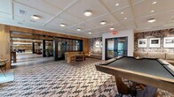 Located at 111 E Broad St in Falls Church, Virginia, this generously sized amenity lounge offers a sophisticated and inviting atmosphere across a single, expansive floor. The space is defined by a striking mosaic tile floor in a multi-toned diamond pattern that flows seamlessly throughout, lending visual continuity and a refined aesthetic to every zone.

Warm natural wood wall paneling and black-framed steel-and-glass partitions create elegant architectural divisions between activity areas without closing off the open feel of the space. A full billiards table anchors one section of the lounge, accompanied by a foosball table positioned near floor-to-ceiling windows that draw in abundant natural light and views of the surrounding streetscape.

A sleek bar area features dark cabinetry, marble-like countertops, and bar seating, accented by dramatic botanical wallcovering as a backdrop. Multiple curated seating vignettes—furnished with mid-century-inspired chairs, sofas, and accent tables—offer relaxed gathering spots throughout. A dedicated reading or library nook with built-in shelving adds a quiet retreat.

The coffered white ceilings provide a clean, classic contrast to the warm wood tones below, while potted tropical plants and framed artwork contribute to the polished yet welcoming ambiance. This richly appointed lounge is designed for both social engagement and quiet relaxation.