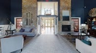 This well-appointed single-floor community space combines nautical-inspired design with refined comfort throughout its interconnected areas. The grand great room serves as the centerpiece, boasting dramatically soaring ceilings clad in natural stone, a striking stone fireplace, and rich navy blue walls accented by crisp white wainscoting. Decorative oars and a model sailboat nod to a coastal aesthetic, while large windows and French doors flood the space with natural light and provide direct access to an outdoor balcony. Multiple seating arrangements feature elegant white armchairs, plush sofas, and warm wood accent tables, creating inviting conversation areas. A dedicated dining nook offers a granite-topped round table surrounded by woven rattan chairs, ideal for casual gatherings. The fully equipped kitchen is a standout feature, showcasing rich cherry wood cabinetry, expansive granite countertops, a central island with a built-in gas cooktop, a stainless steel range hood, double wall ovens, and a breakfast bar with seating. Tile flooring runs throughout the kitchen, while patterned carpeting lends warmth to the lounge areas. Together, the spaces deliver a cohesive, upscale ambiance perfectly suited for entertaining and community gatherings.