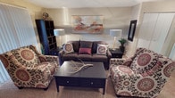 This single-floor space centers around a thoughtfully designed living room that balances comfort and style. The room features a deep charcoal-brown sofa adorned with patterned and solid accent pillows, flanked by two generously sized armchairs upholstered in a bold floral print with red and cream tones. A dark-finished coffee table with drawer storage serves as the focal point of the seating arrangement, topped with a decorative driftwood branch that adds a natural element to the space.

Two matching table lamps with light blue shades provide warm ambient lighting from dark-stained end tables positioned on either side of the sofa. A large abstract canvas in warm reds, oranges, and blues anchors the wall above the sofa, adding artistic character. A tall black bookcase unit offers vertical storage and display space along one wall, while a framed mirror on the opposite side enhances the sense of depth. Neutral beige carpeting and light-toned walls create a cohesive backdrop, lending the overall space a warm, welcoming ambiance suitable for everyday living and entertaining.