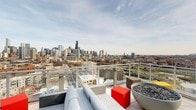 This exceptional rooftop terrace delivers one of the most commanding views of the Chicago skyline available, with iconic landmarks such as the Willis Tower rising prominently against the horizon. The outdoor space is thoughtfully designed for both relaxation and entertainment, featuring contemporary lounge furniture including a plush gray sectional sofa paired with a bold red accent cube for casual seating. A large bowl-style fire pit filled with lava rocks serves as a striking centerpiece, providing warmth and ambiance during cooler evenings. The terrace is enclosed by a sleek glass and metal railing system that preserves unobstructed sightlines across the city's urban landscape, stretching from the dense downtown core to the surrounding neighborhoods below. The open-air design allows for abundant natural light throughout the day, while the elevated vantage point captures sweeping views of Chicago's streets, infrastructure, and architectural diversity. The clean, modern aesthetic of the terrace complements the dynamic cityscape backdrop, creating a sophisticated outdoor retreat ideal for gatherings or quiet reflection high above the city.