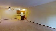 This single-floor space offers an open-concept layout that connects the living area and kitchen in a cohesive, functional design. The living area is finished with wall-to-wall carpet in a neutral beige tone, providing a comfortable and quiet underfoot feel. Crisp white walls and standard-height ceilings keep the space feeling clean and bright.

The kitchen is positioned at the rear of the open floor plan and is defined by a peninsula-style breakfast bar with a dark countertop, creating a natural visual boundary between the two areas. Warm honey-toned wood cabinetry lines the upper and lower walls, complemented by white appliances including a range and over-the-range microwave. A set of pendant lights hangs above the kitchen area, adding focused task lighting and a subtle decorative element. The kitchen floor transitions to a hard surface, contrasting with the carpeted living space.

A ceiling fan is partially visible in the living area, contributing to air circulation. Neutral baseboards frame the walls throughout, and a visible HVAC vent in the ceiling ensures climate control. Overall, the space presents a straightforward, functional layout suited for everyday living.