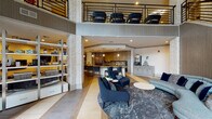Located at 1300 Tribute Center Dr in Raleigh, NC, this upscale communal amenity space is thoughtfully designed to blend comfort, function, and contemporary style. The main lounge area makes an immediate impression with soaring double-height ceilings and an open mezzanine level above, finished with a sleek metal railing that overlooks the activity below. Stacked stone columns anchor the space architecturally, adding warmth and texture that contrasts beautifully with the modern finishes throughout.

The lounge is organized into distinct seating zones, including a plush velvet sectional sofa paired with a solid wood coffee table, positioned beside floor-to-ceiling windows that invite abundant natural light and views of the surrounding greenery. A linear wall-mounted fireplace serves as a sophisticated focal point in one seating area, enhancing the space's inviting ambiance. Patterned tile and area rugs define each zone, adding visual interest underfoot.

The fully equipped kitchen is a standout feature, outfitted with two-tone cabinetry, quartz countertops, a large island with bar seating, stainless steel appliances, a tile backsplash, and pendant lighting. Adjacent to the kitchen, a dedicated beverage and service station provides added convenience. The overall design palette of navy, gold, gray, and warm neutrals creates a cohesive, refined atmosphere throughout.