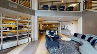 Located at 1300 Tribute Center Dr in Raleigh, NC, this upscale communal amenity space is thoughtfully designed to blend comfort, function, and contemporary style. The main lounge area makes an immediate impression with soaring double-height ceilings and an open mezzanine level above, finished with a sleek metal railing that overlooks the activity below. Stacked stone columns anchor the space architecturally, adding warmth and texture that contrasts beautifully with the modern finishes throughout.

The lounge is organized into distinct seating zones, including a plush velvet sectional sofa paired with a solid wood coffee table, positioned beside floor-to-ceiling windows that invite abundant natural light and views of the surrounding greenery. A linear wall-mounted fireplace serves as a sophisticated focal point in one seating area, enhancing the space's inviting ambiance. Patterned tile and area rugs define each zone, adding visual interest underfoot.

The fully equipped kitchen is a standout feature, outfitted with two-tone cabinetry, quartz countertops, a large island with bar seating, stainless steel appliances, a tile backsplash, and pendant lighting. Adjacent to the kitchen, a dedicated beverage and service station provides added convenience. The overall design palette of navy, gold, gray, and warm neutrals creates a cohesive, refined atmosphere throughout.