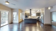 Located in Dallas, this single-floor space showcases a thoughtfully designed open-concept layout that seamlessly blends style and functionality. The kitchen serves as the centerpiece, featuring sleek gray cabinetry, a large center island with a stone countertop, and a suite of stainless steel appliances including a double wall oven, a built-in microwave, and a full-size refrigerator. A statement stainless steel range hood anchors the cooking area, venting above a gas cooktop set into the island. A mosaic tile backsplash adds texture and visual interest behind the countertops. Recessed lighting and pendant fixtures provide ample illumination throughout the space. Rich, dark hardwood flooring extends across the open floor plan, offering warmth and contrast against the light-toned walls and cabinetry. Multiple windows allow natural light to filter in, creating a bright and welcoming ambiance. The generous open layout between the kitchen and adjoining living area provides a flexible and expansive feel, ideal for both everyday living and entertaining. Overall, the space presents a clean, modern aesthetic with high-quality finishes and a well-considered design throughout.