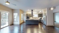 Located in Dallas, this single-floor space showcases a thoughtfully designed open-concept layout that seamlessly blends style and functionality. The kitchen serves as the centerpiece, featuring sleek gray cabinetry, a large center island with a stone countertop, and a suite of stainless steel appliances including a double wall oven, a built-in microwave, and a full-size refrigerator. A statement stainless steel range hood anchors the cooking area, venting above a gas cooktop set into the island. A mosaic tile backsplash adds texture and visual interest behind the countertops. Recessed lighting and pendant fixtures provide ample illumination throughout the space. Rich, dark hardwood flooring extends across the open floor plan, offering warmth and contrast against the light-toned walls and cabinetry. Multiple windows allow natural light to filter in, creating a bright and welcoming ambiance. The generous open layout between the kitchen and adjoining living area provides a flexible and expansive feel, ideal for both everyday living and entertaining. Overall, the space presents a clean, modern aesthetic with high-quality finishes and a well-considered design throughout.