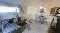 This three-floor space offers a thoughtfully designed layout that maximizes both function and style. The main living area showcases a combined living and dining space bathed in natural light from well-placed windows. Neutral-toned walls provide a warm, inviting backdrop, while plush wall-to-wall carpeting adds comfort underfoot.

The living area is anchored by a comfortable seating arrangement, complemented by decorative wall art that adds an artistic focal point to the space. A set of modern nesting tables serves as a versatile coffee table solution, enhancing the room's contemporary aesthetic.

Adjacent to the living area, the dining space features a raised pub-style table with matching bar stools, illuminated by a stylish chandelier overhead. A decorative mirror on the dining area wall adds depth and visual interest. The open-concept arrangement between the living and dining areas creates a seamless flow, making the space feel expansive.

A staircase is prominently visible, connecting the multiple floors and contributing to the home's sense of vertical space. The overall ambiance is modern and inviting, with carefully chosen decorative elements that enhance the neutral color palette throughout.