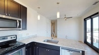 This single-floor residence showcases a clean, modern design with an open-concept layout that maximizes the sense of space and flow. The kitchen is a standout feature, equipped with rich dark cabinetry that contrasts elegantly against light granite countertops. A deep stainless steel sink is set into the granite island, which also serves as a natural divider between the kitchen and living areas. Stainless steel appliances, including a dishwasher, add a polished, contemporary touch. Two cylindrical pendant lights hang above the kitchen workspace, providing focused task lighting while contributing to the modern aesthetic. The living area beyond the kitchen benefits from a ceiling fan with a sleek, brushed nickel finish, ensuring year-round comfort. Crisp white walls and smooth ceilings amplify the brightness throughout the space, creating an airy and inviting atmosphere. A hallway visible from the kitchen leads to additional rooms, hinting at a thoughtfully designed floor plan. The overall ambiance is fresh, clean, and move-in ready, offering a well-appointed environment suited for modern living.