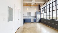 This single-floor loft showcases a striking blend of industrial heritage and contemporary design. Polished concrete floors run throughout the open layout, reflecting natural light that pours in through expansive, multi-pane factory-style windows framed in dark steel. The painted brick walls add raw character and depth to the space, while soaring ceilings with exposed ductwork, cofferred concrete slab details, and vivid yellow-orange overhead pipes reinforce the unit's industrial roots.

The open living area is generous and airy, anchored by the dramatic floor-to-ceiling windows that offer sweeping city views. Globe pendant lights hang from the high ceilings, providing warm ambient illumination.

The kitchen is thoughtfully designed with a two-tone cabinet scheme — crisp white uppers with open shelving paired with slate-blue lower cabinets — set against a textured tile backsplash. Stainless steel appliances, including a range with over-the-range microwave, dishwasher, and refrigerator, complete the functional and stylish cooking space with clean white countertops tying it all together.

The bathroom continues the clean, modern aesthetic with white cabinetry, a full tub, and polished concrete flooring. The overall ambiance of the space is modern industrial — open, light-filled, and full of architectural character.